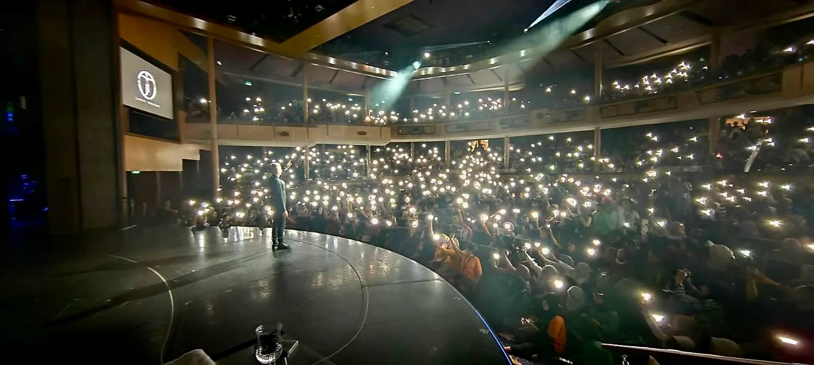 John Taylor performing on stage before a theatre audience raising phone lights